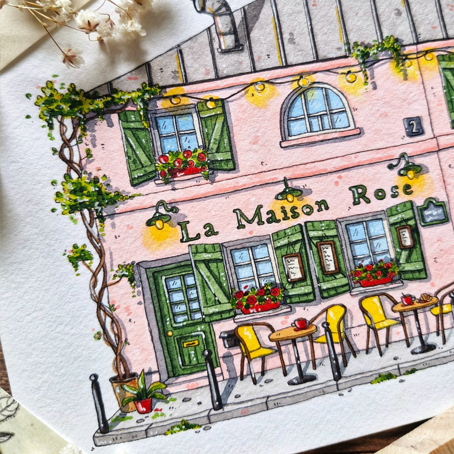 Illustration of a quaint street scene with 'La Maison Rose' building, flowers, and chairs.