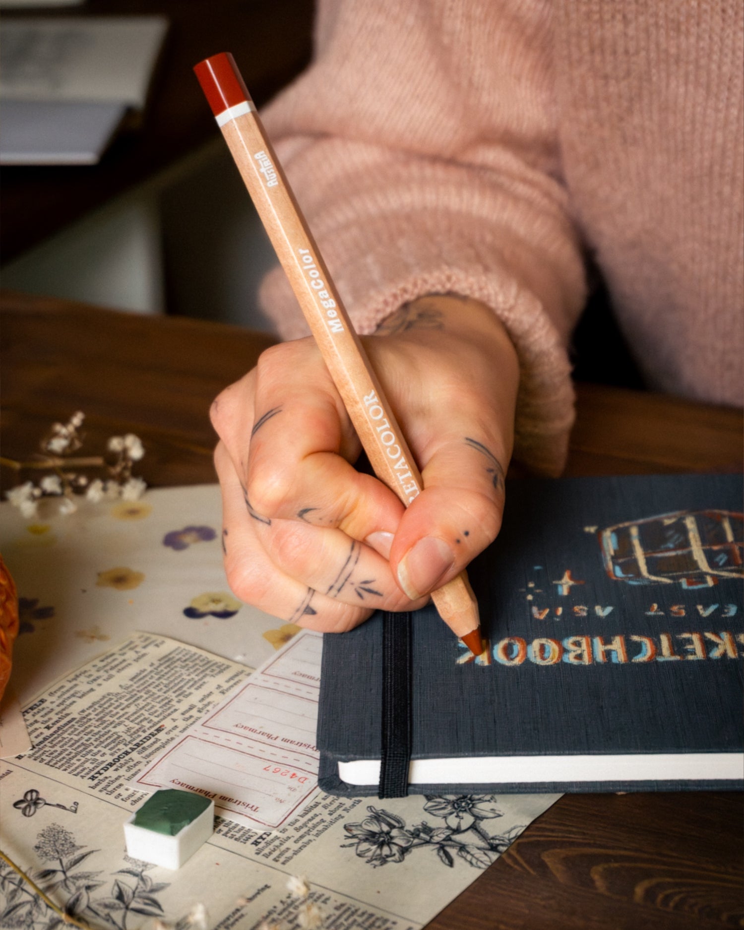 Hand holding a pencil over a notebook with a decorative cover on a wooden surface.