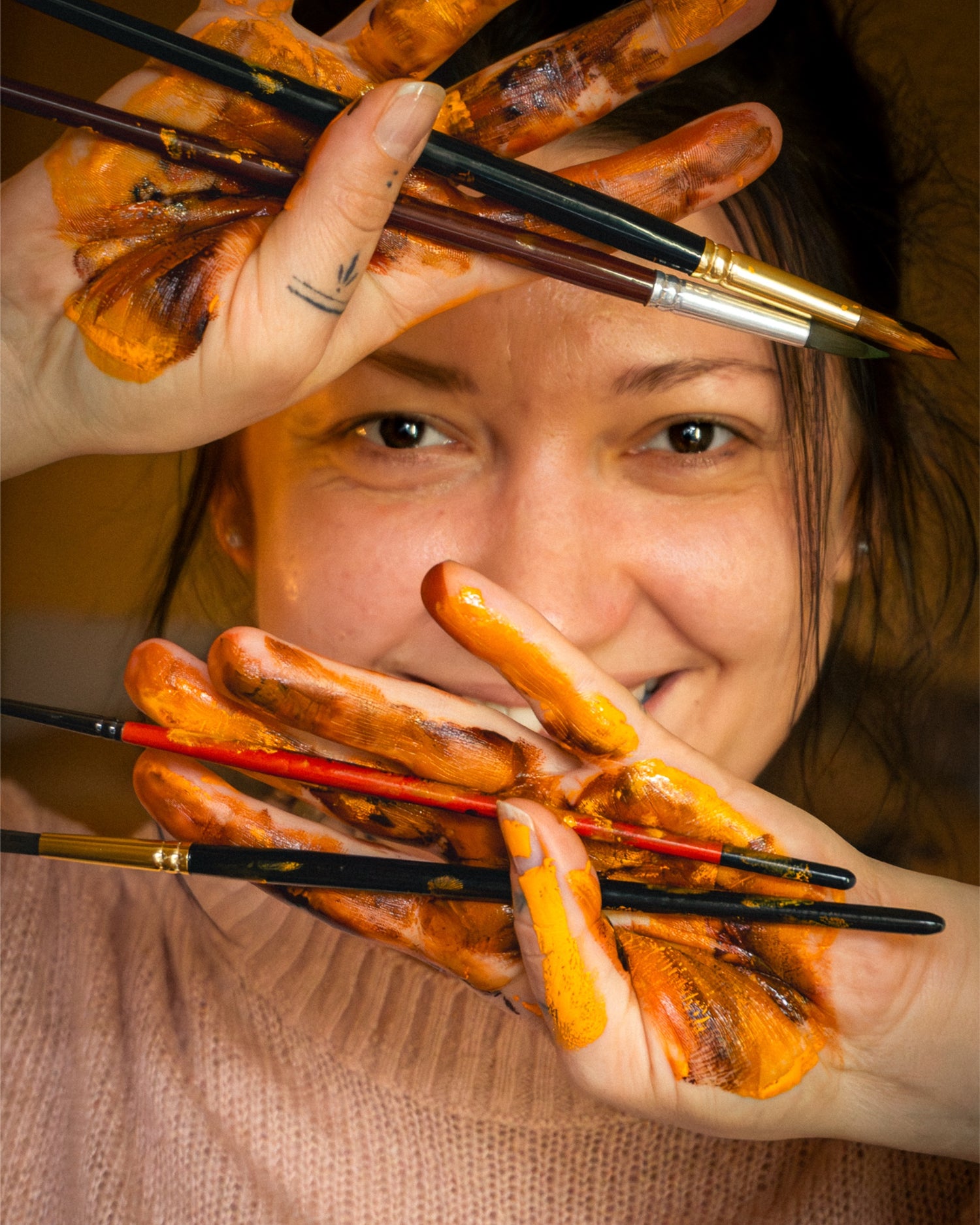 Person with orange paint on hands holding brushes, with a blurred background