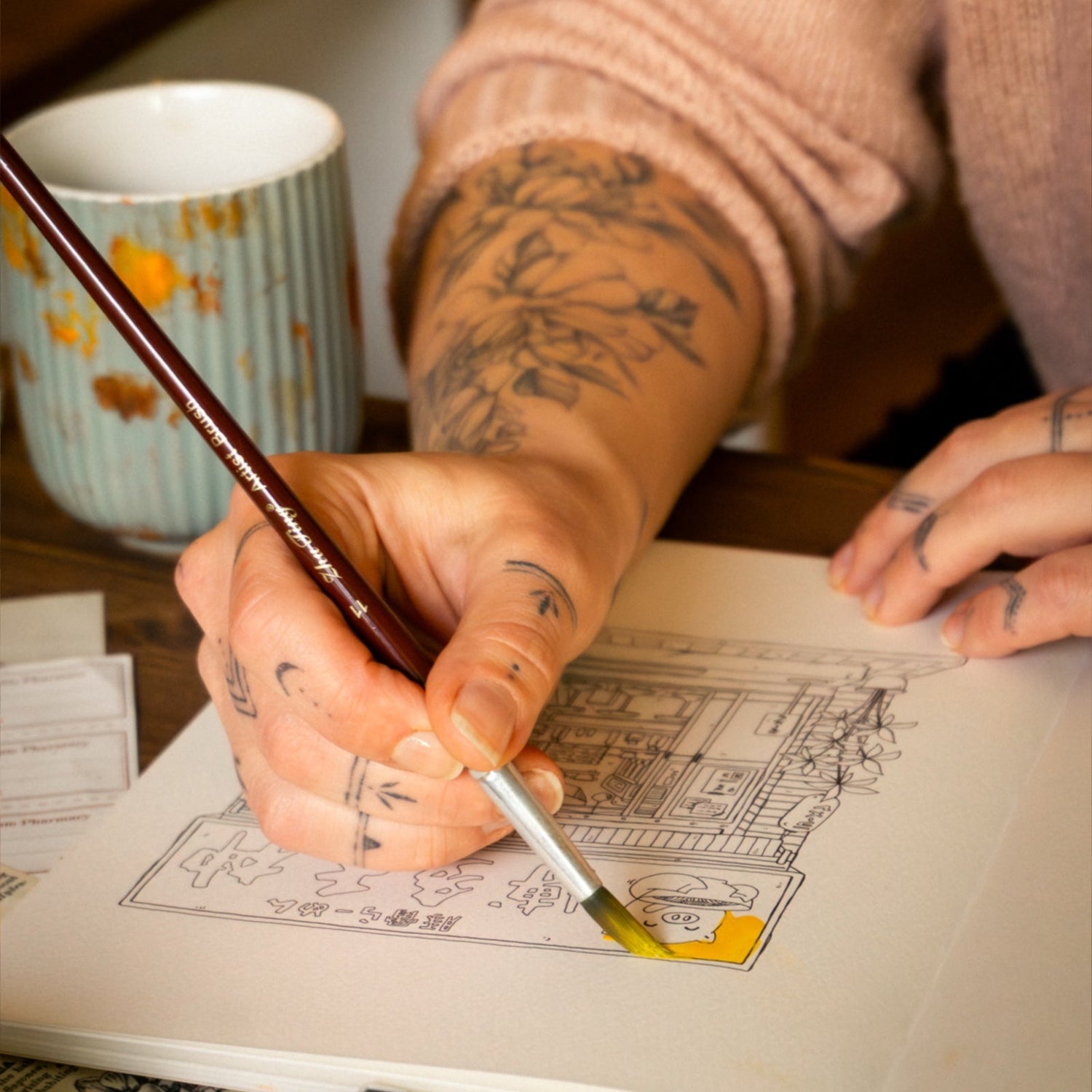 Person with tattoos painting a detailed illustration in a sketchbook with a mug in the background.